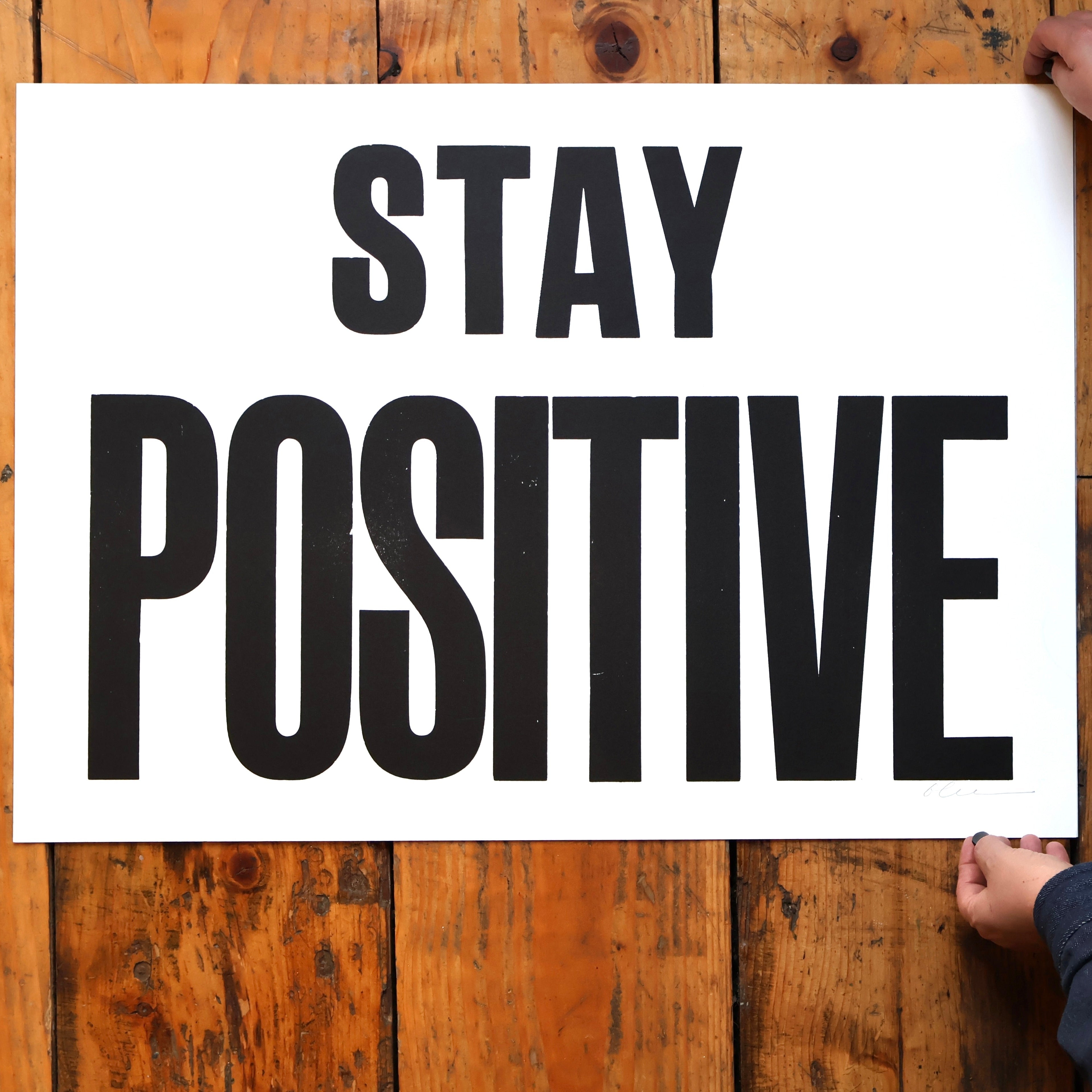Person holding a sign with 'Stay Positive' text on a wooden surface