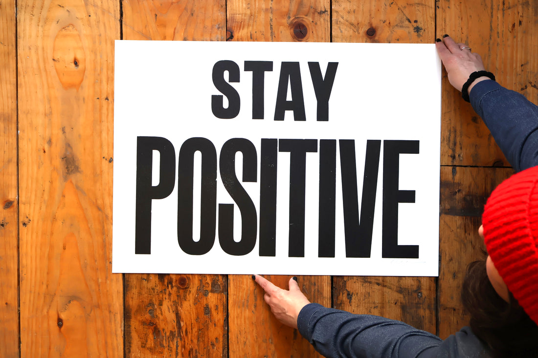 Person holding a sign with 'Stay Positive' text on a wooden surface