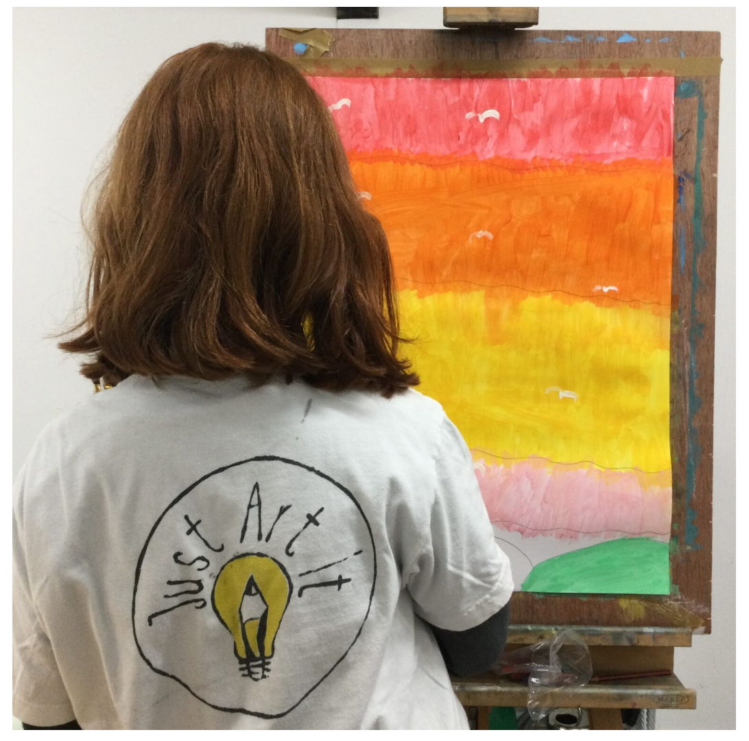Back of child painting on paper with an easel, their painting is red, orange yellow pink and green. They are wearing a Just Art it Galway t-shirt.
