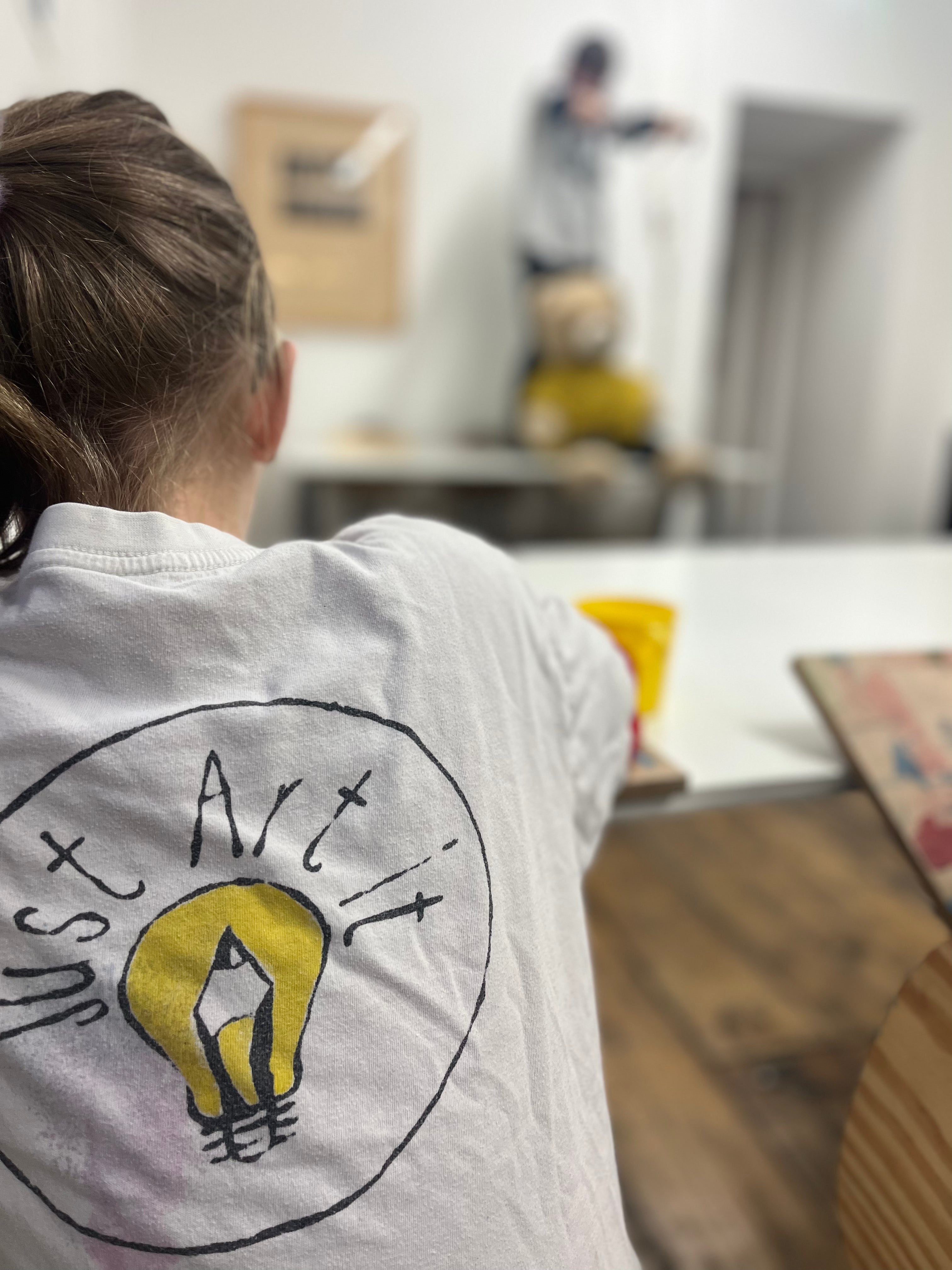Child wearing a shirt with 'Just Art It' Original Logo in a life drawing setting, drawing with their pals posing with our teddy bear Bob