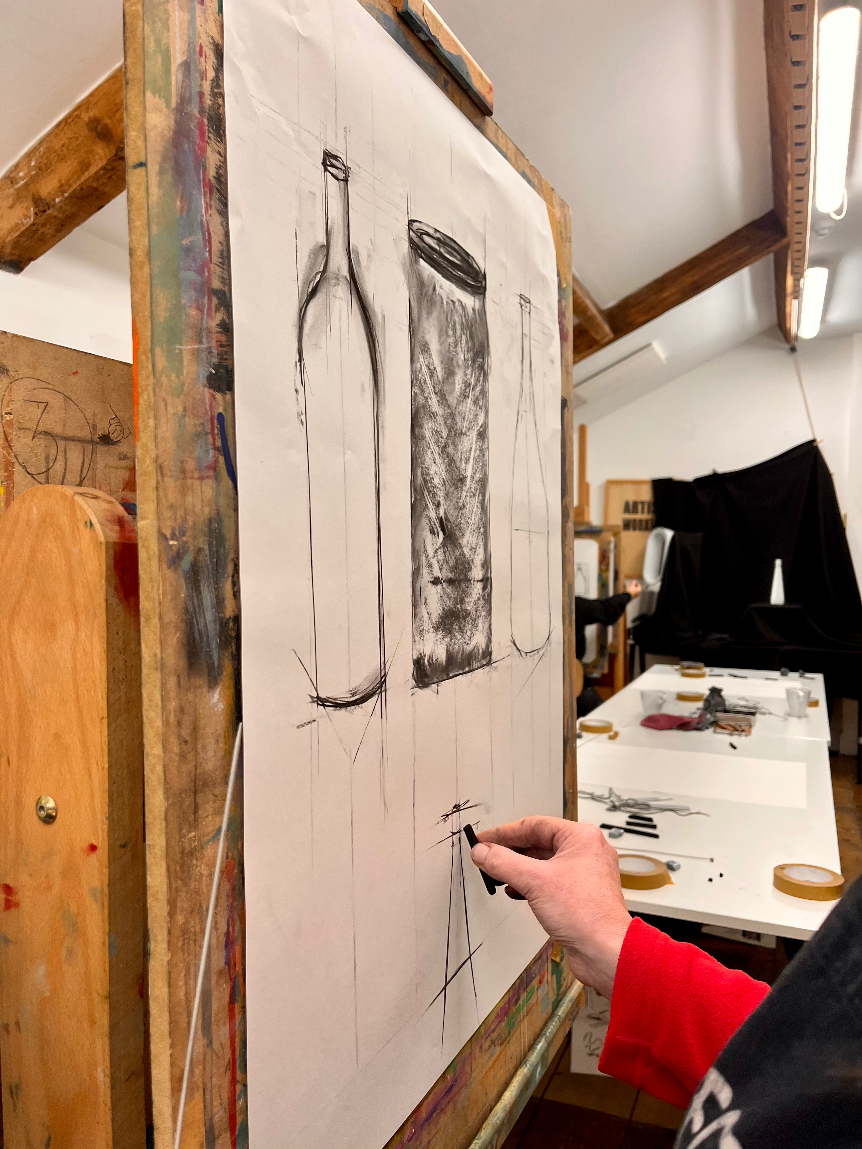 Person drawing on a large sheet of paper with a bottle and rectangular object sketch.