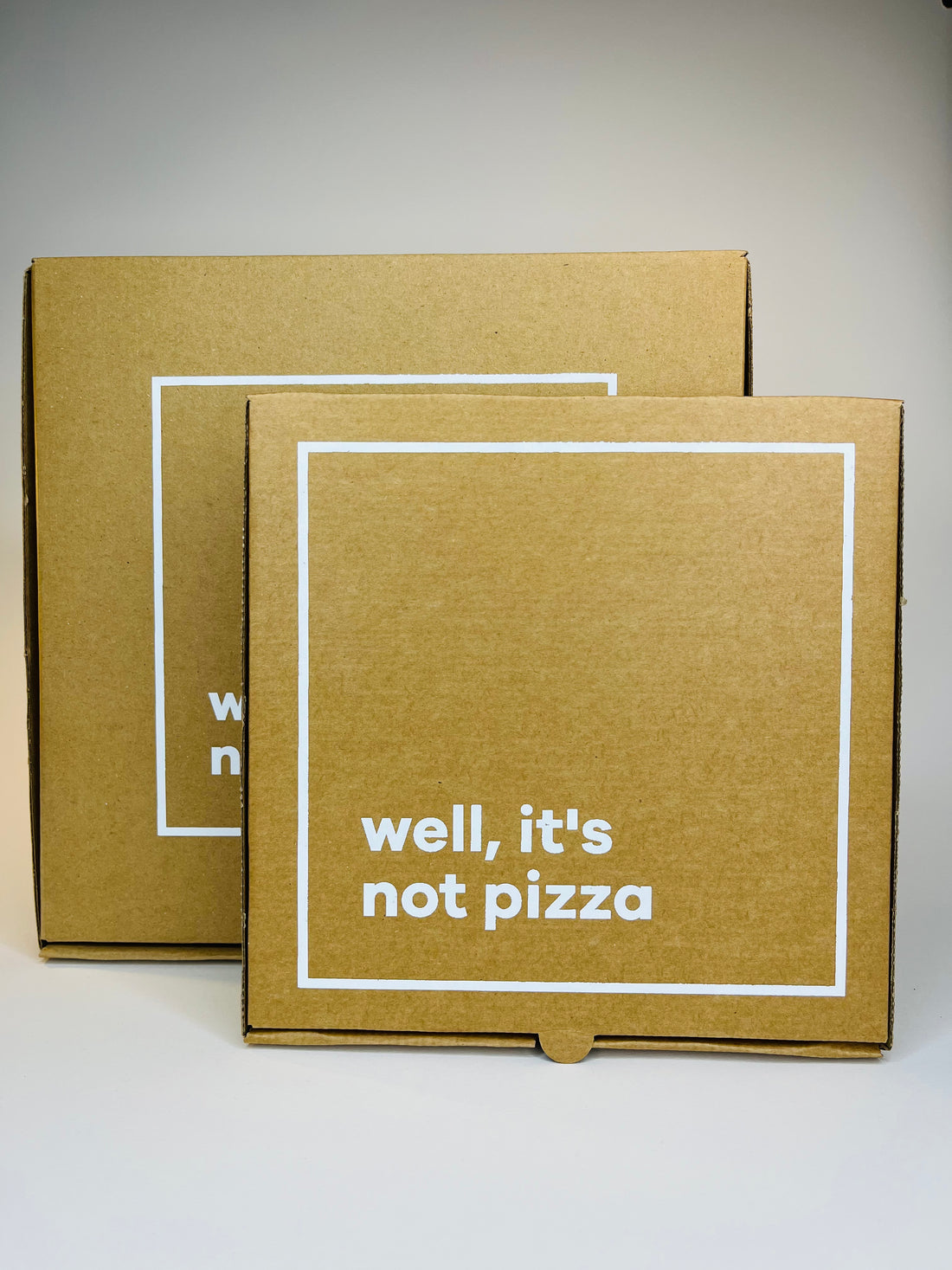 Two brown cardboard boxes with white square borders, one displaying "well, it's not pizza" in white text.