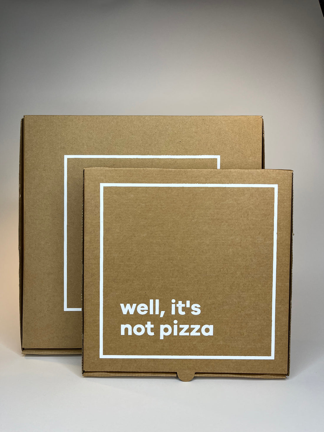 Two brown cardboard boxes with white square borders, one displaying "well, it's not pizza" in white text.