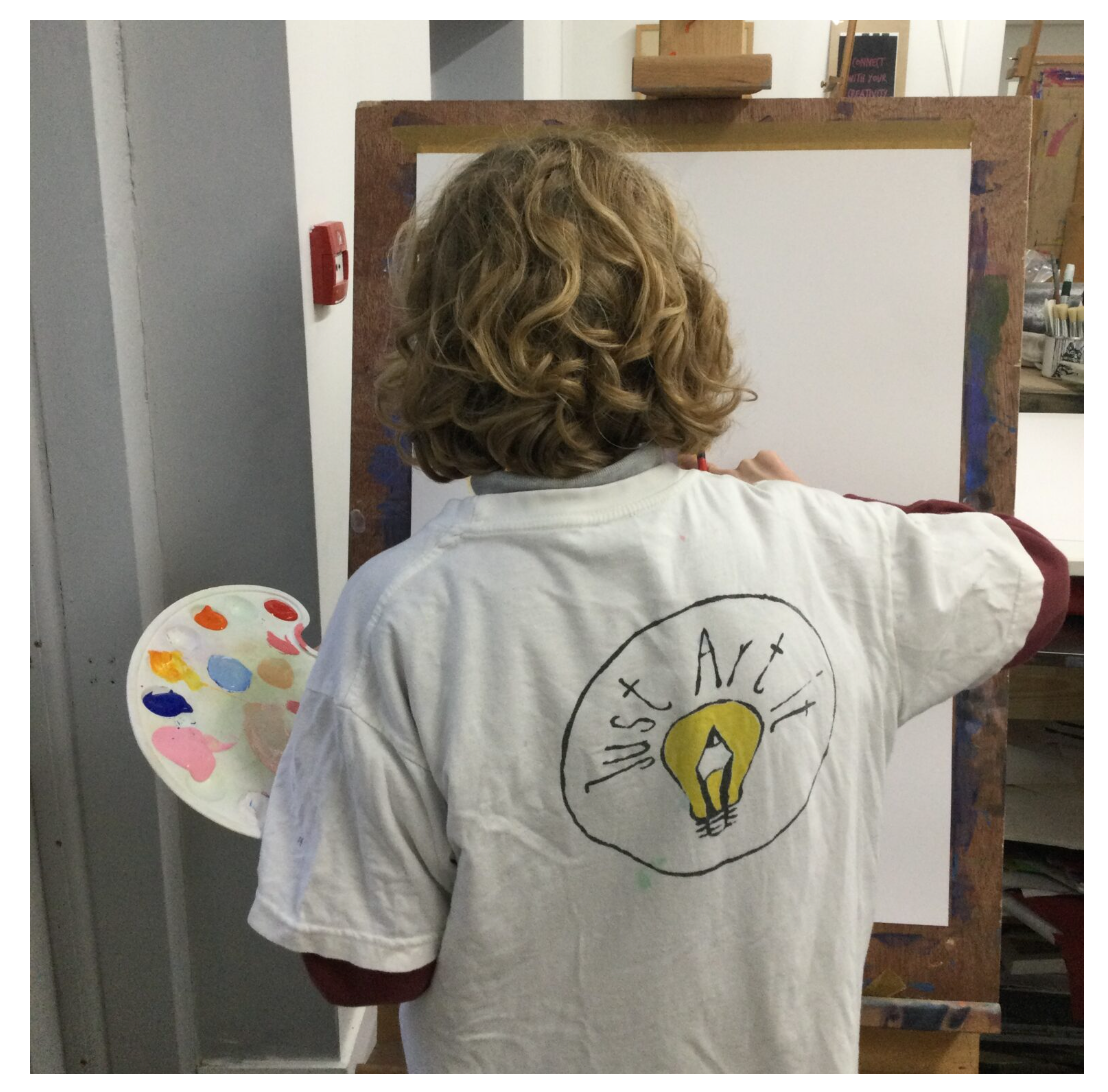 A child painting in an art class, seen from behind, with a paint palette in hand, standing in front of an easel.
