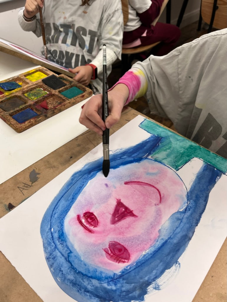 Child painting a self-portrait with pink face and blue hair on paper, using a ArtGraf water soluble pigments, palette and brush.