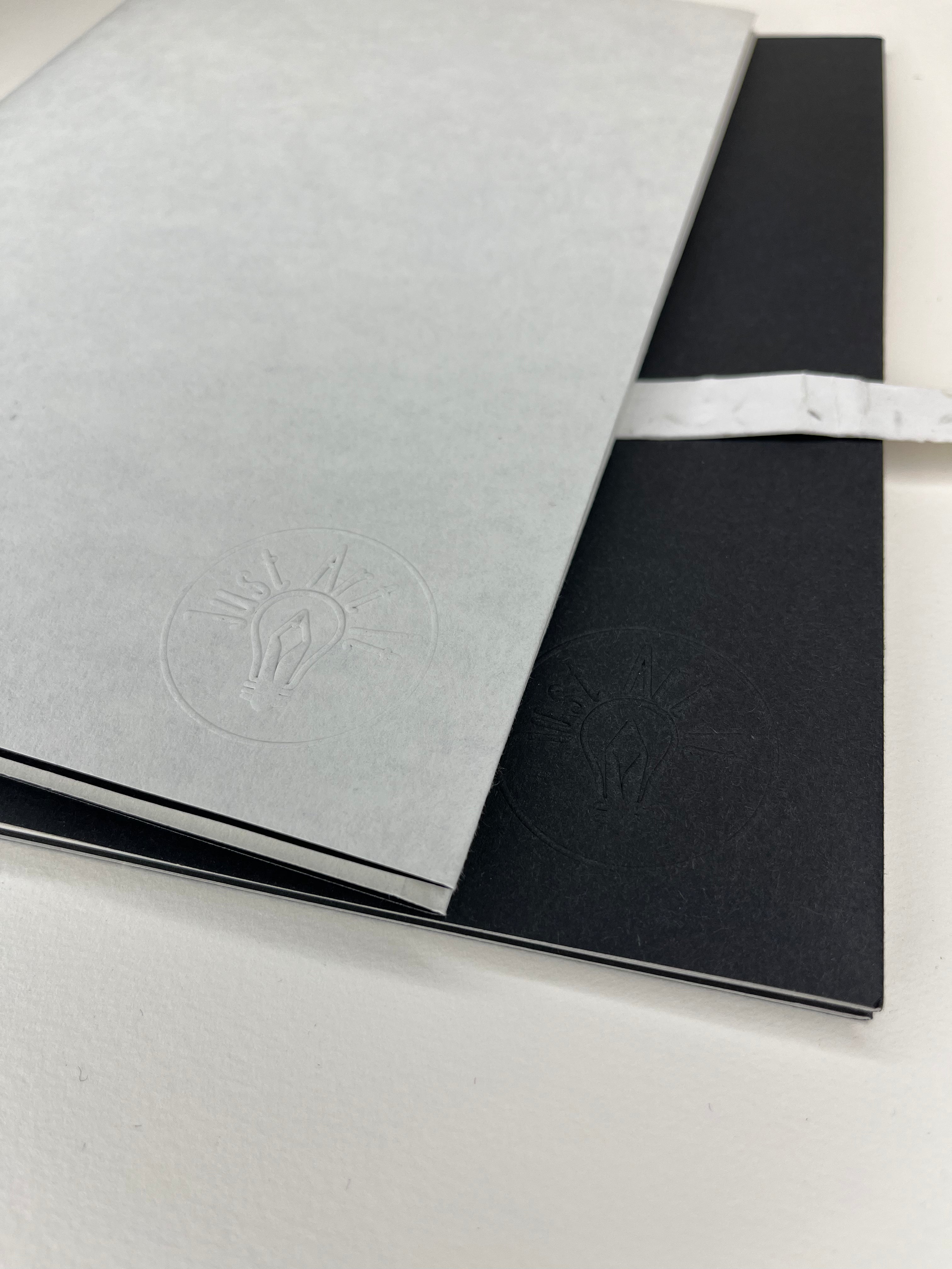 Two notebooks, one light gray and one black, rest on a white surface. Both feature an embossed logo with a lightbulb motif. A white textured ribbon wraps around the black notebook.