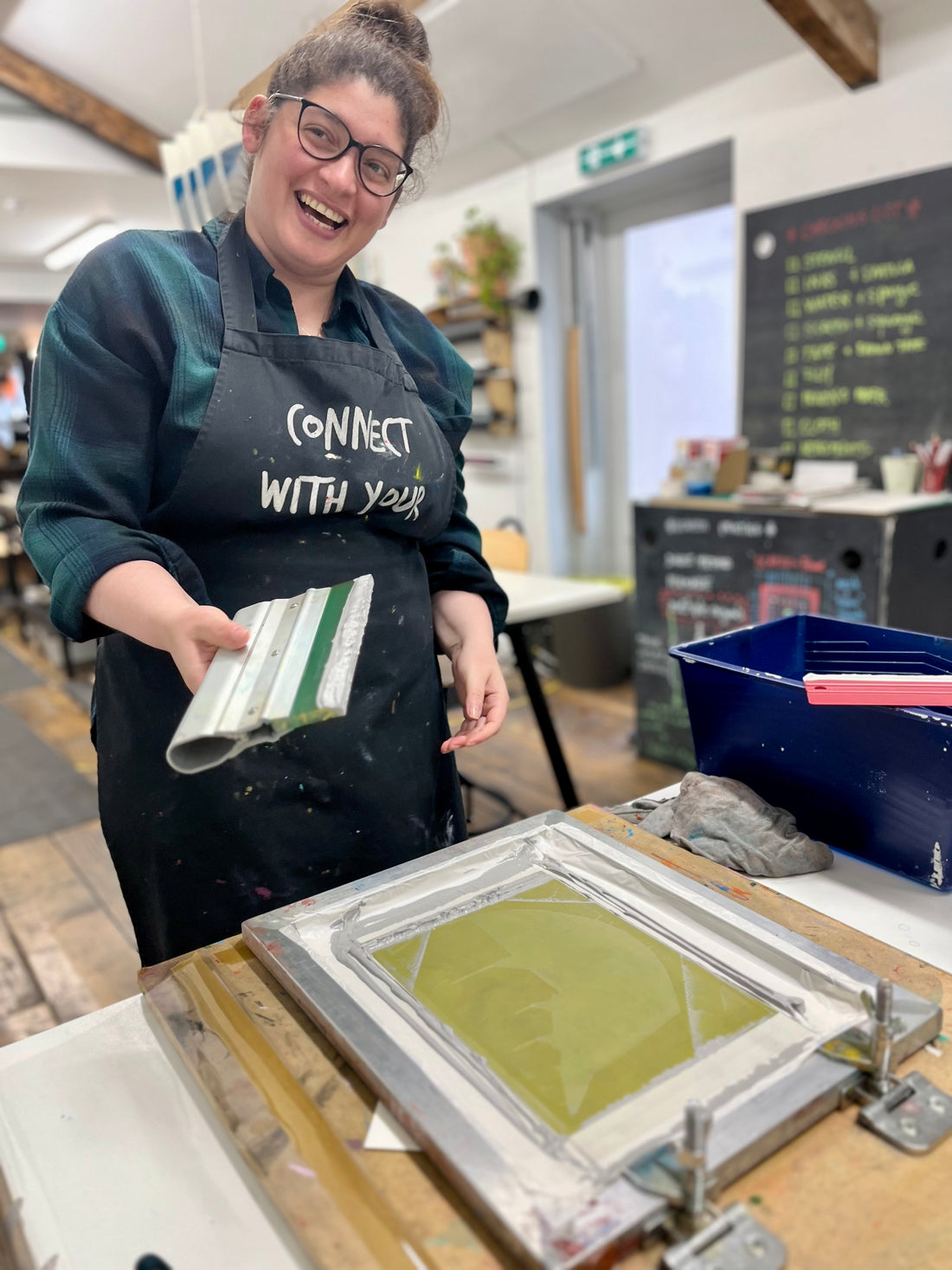 Person wearing a black apron with 'Connect With Your Creativity" text, holding a screen printing frame with green ink.