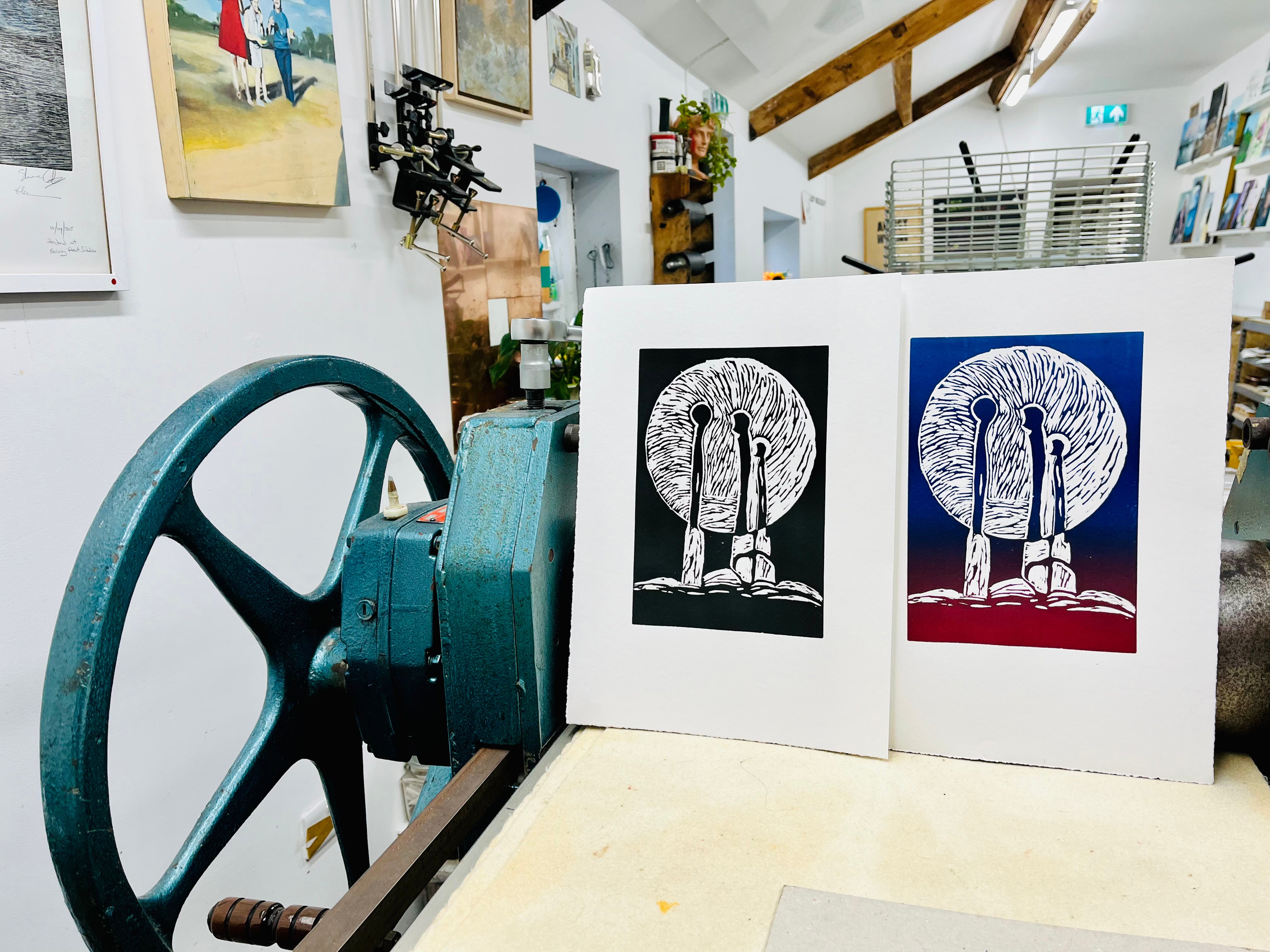 Two prints of a stylized figure in a circular design on a table with a large wheel in the background.