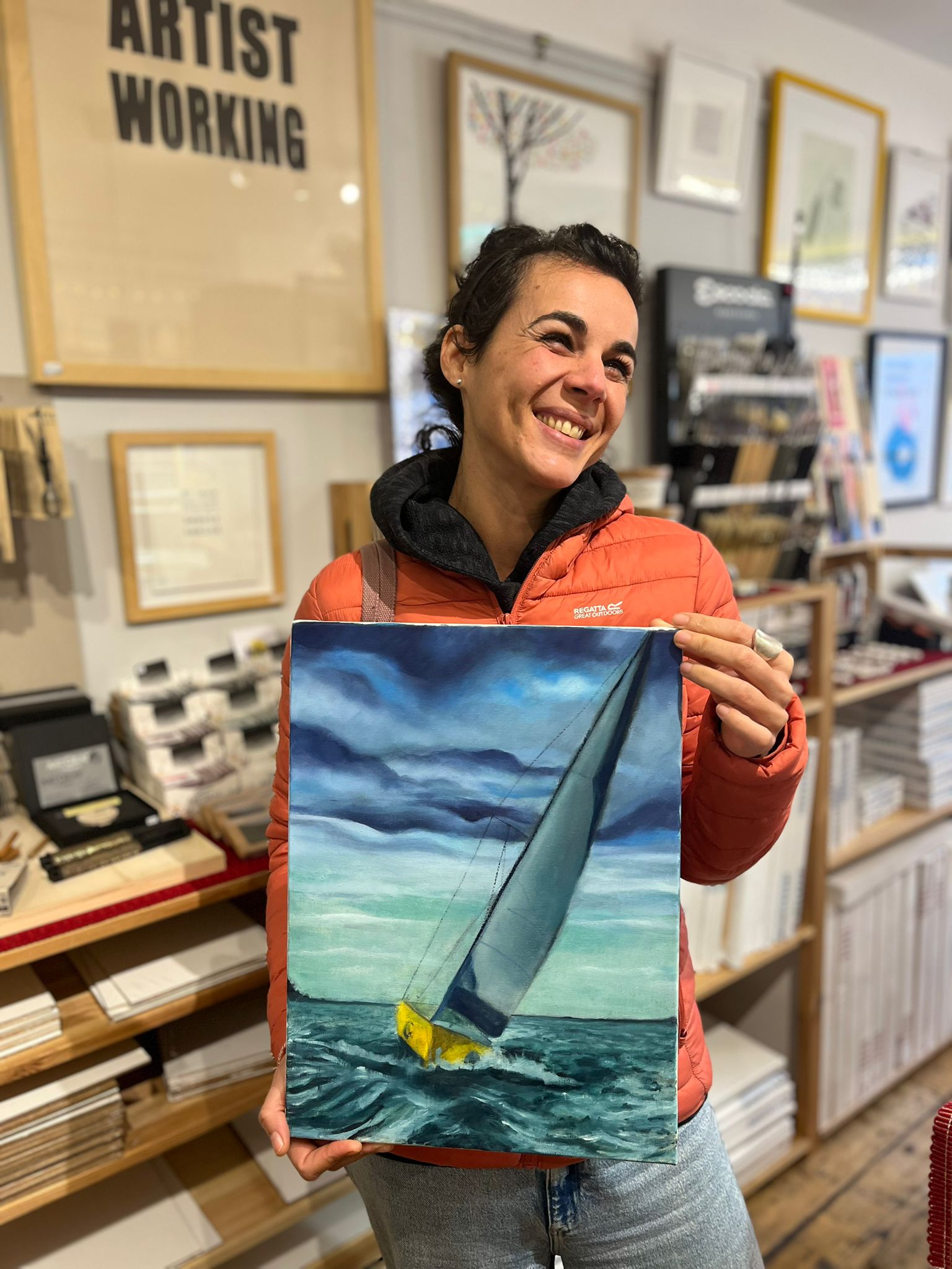 Just Art It' Student holding her sailboat painting in our art shop nearby our Artist Working Print. 
