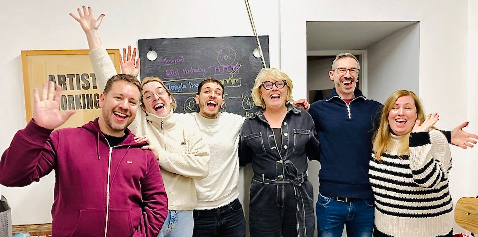 Group of six people posing joyfully in a room with a chalkboard in the background.