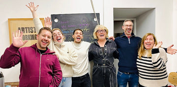 Group of six people posing joyfully in a room with a chalkboard in the background.
